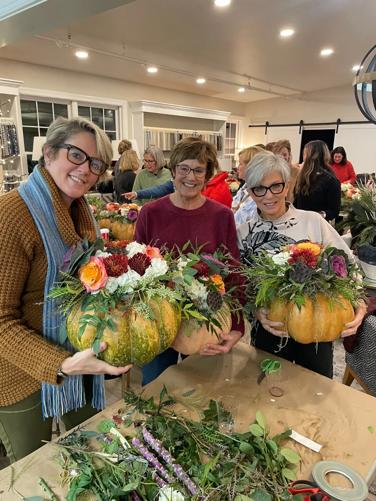 Workshop guests smiling and holding their finished fall floral arrangements in pumpkins