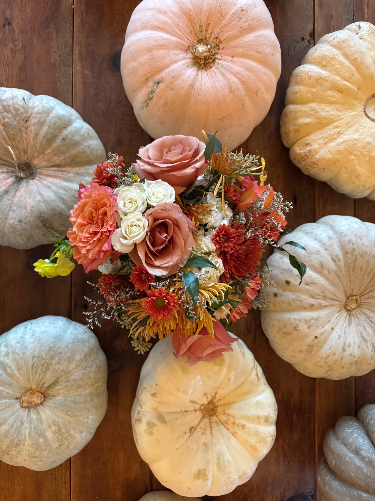 A finished fall centerpiece of roses and mums arranged atop a Cinderella pumpkin, surrounded by heirloom pumpkins